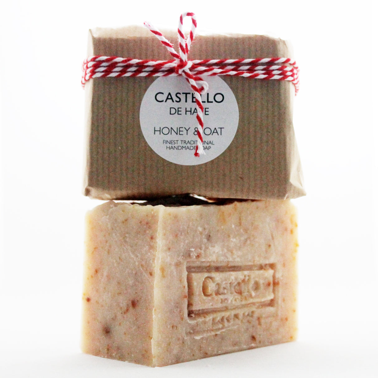 Two bars of soap, the top one wrapped in brown kraft paper with a red and white twine, the bottom one is beige with exfoliating oats visible throughout the texture. Both bars have the 'Castello De Haye' brand name on them.