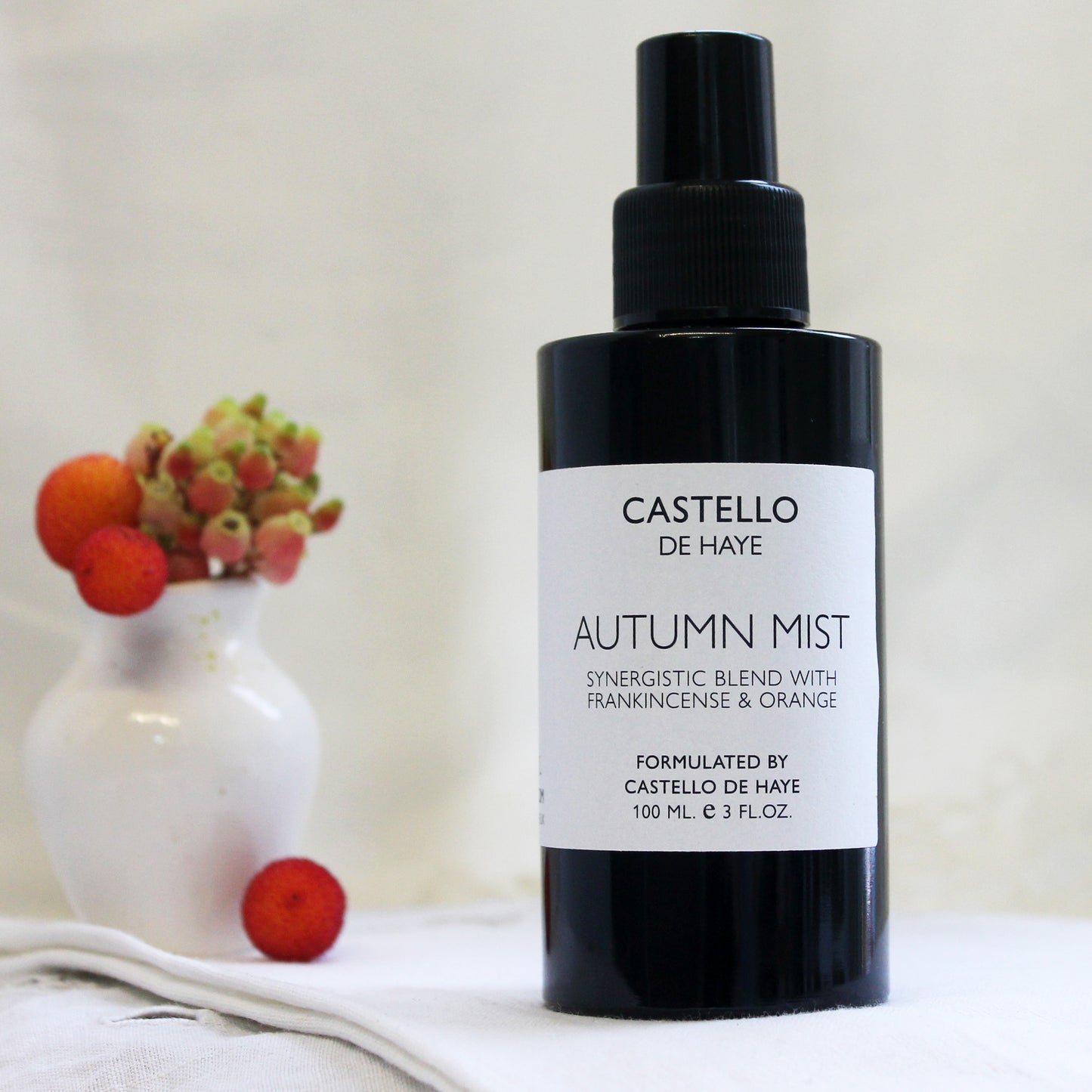 A black spray bottle labeled 'Castello de Haye Autumn Mist' with a white vase containing red flowers in the background.