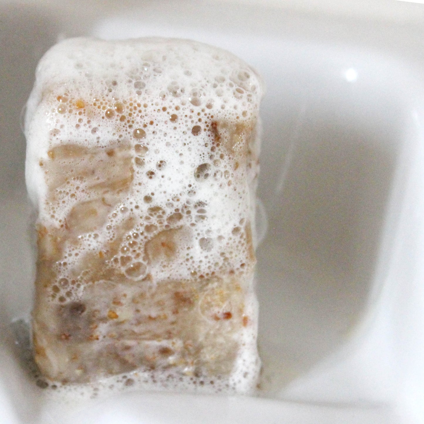 A bar of soap with a creamy oatmeal and honey texture, shown producing soap suds in a bathtub.