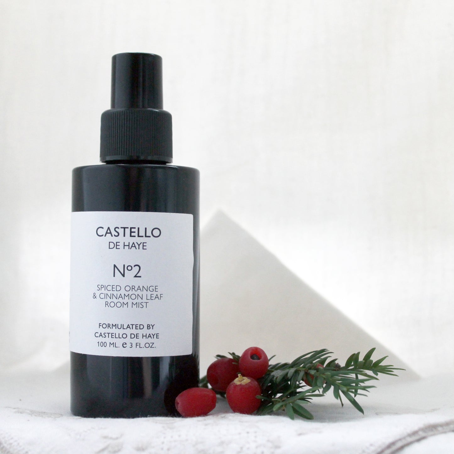 A black spray bottle labeled 'Castello de Haye, No. 2 Spiced Orange & Cinnamon Leaf Room Mist' with white and red text, set against a neutral background with a white cloth and some green leaves and red berries.