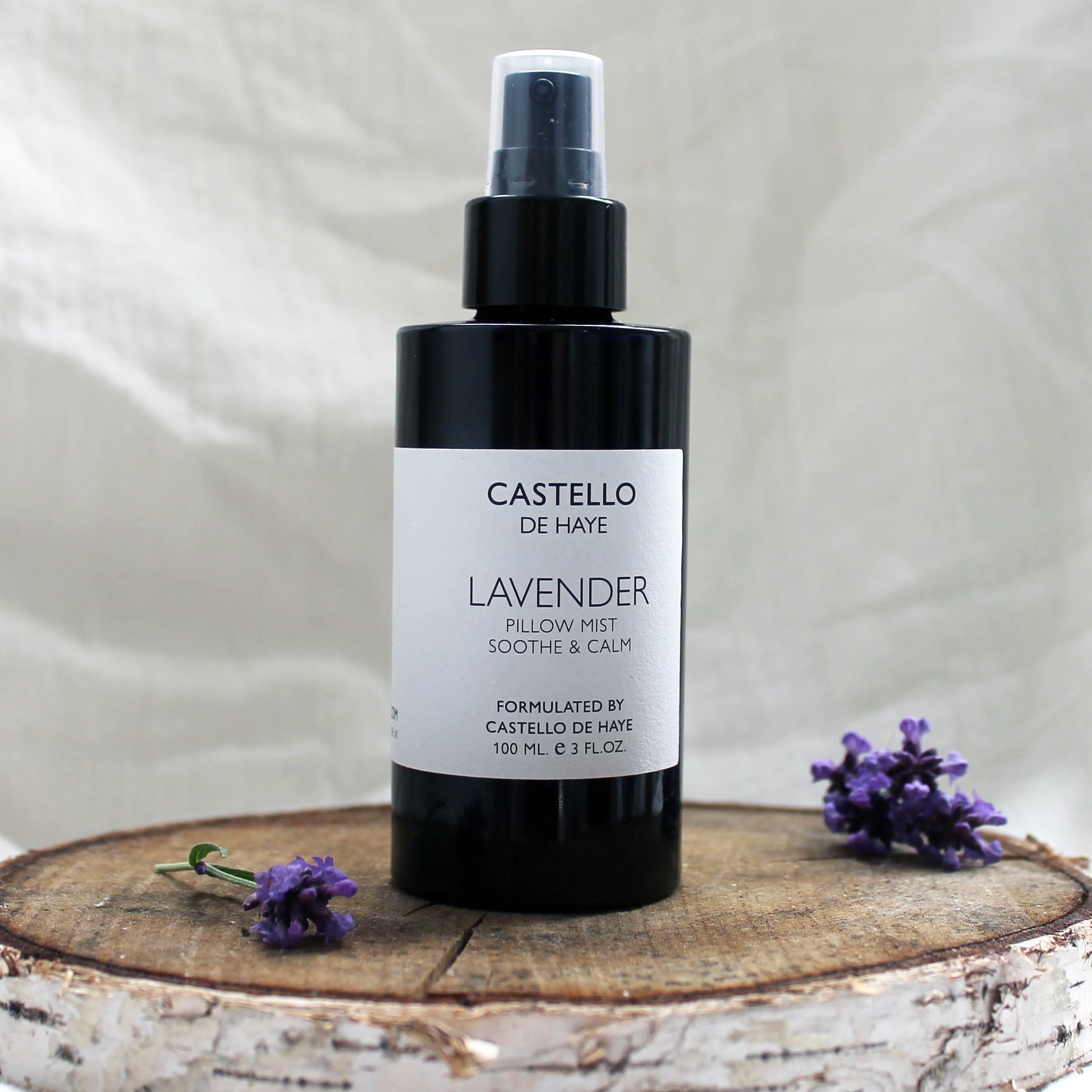 A black bottle of Castello de Haye Lavender Aromatherapy Pillow Mist Spray with a spray nozzle, labeled with the product name and weight of 100ml. The bottle is on a wooden surface with a few sprigs of lavender around it.