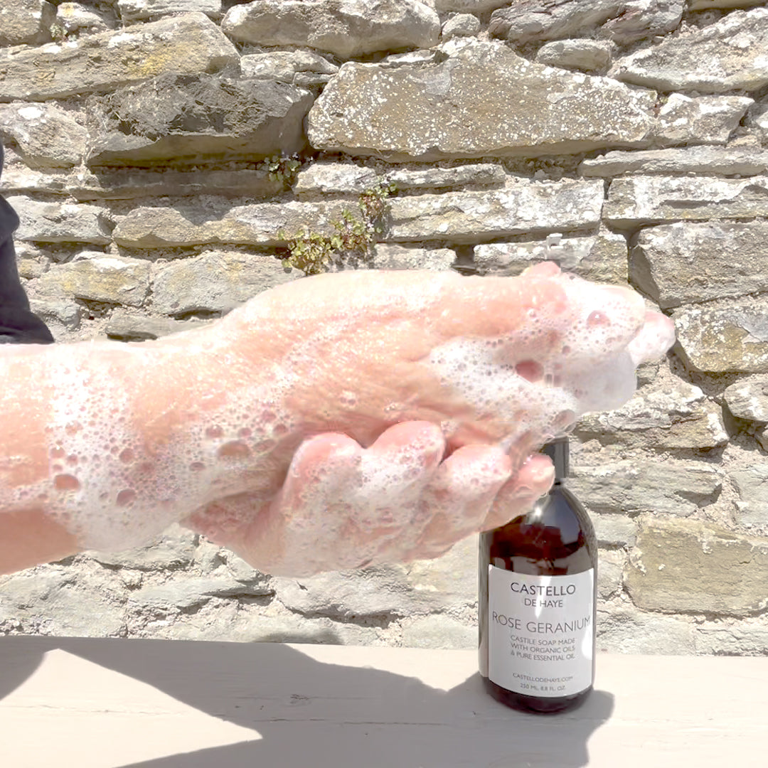 Hand with soap suds holding a bottle of Castello de Haye Rose Geranium soap against a stone wall.