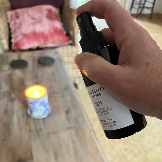 Hand holding No.1 Frankincense & Rose Geranium room mist in a lounge with a lit candle.