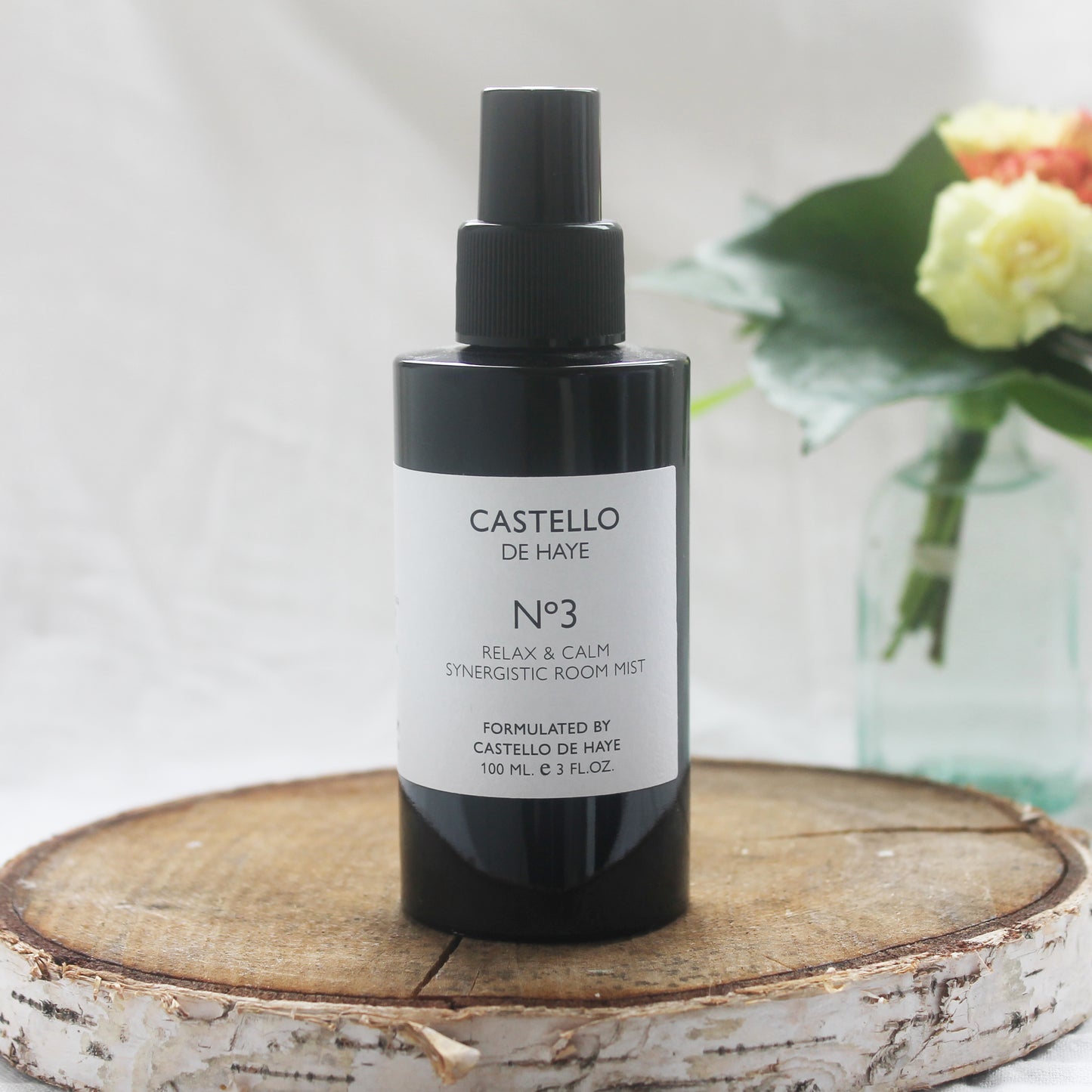 A black spray bottle labeled 'Castello de Haye, No.3 Relax Aromatherapy Room Mist Spray' with a white and purple label, on a wooden surface with a floral arrangement in the background.