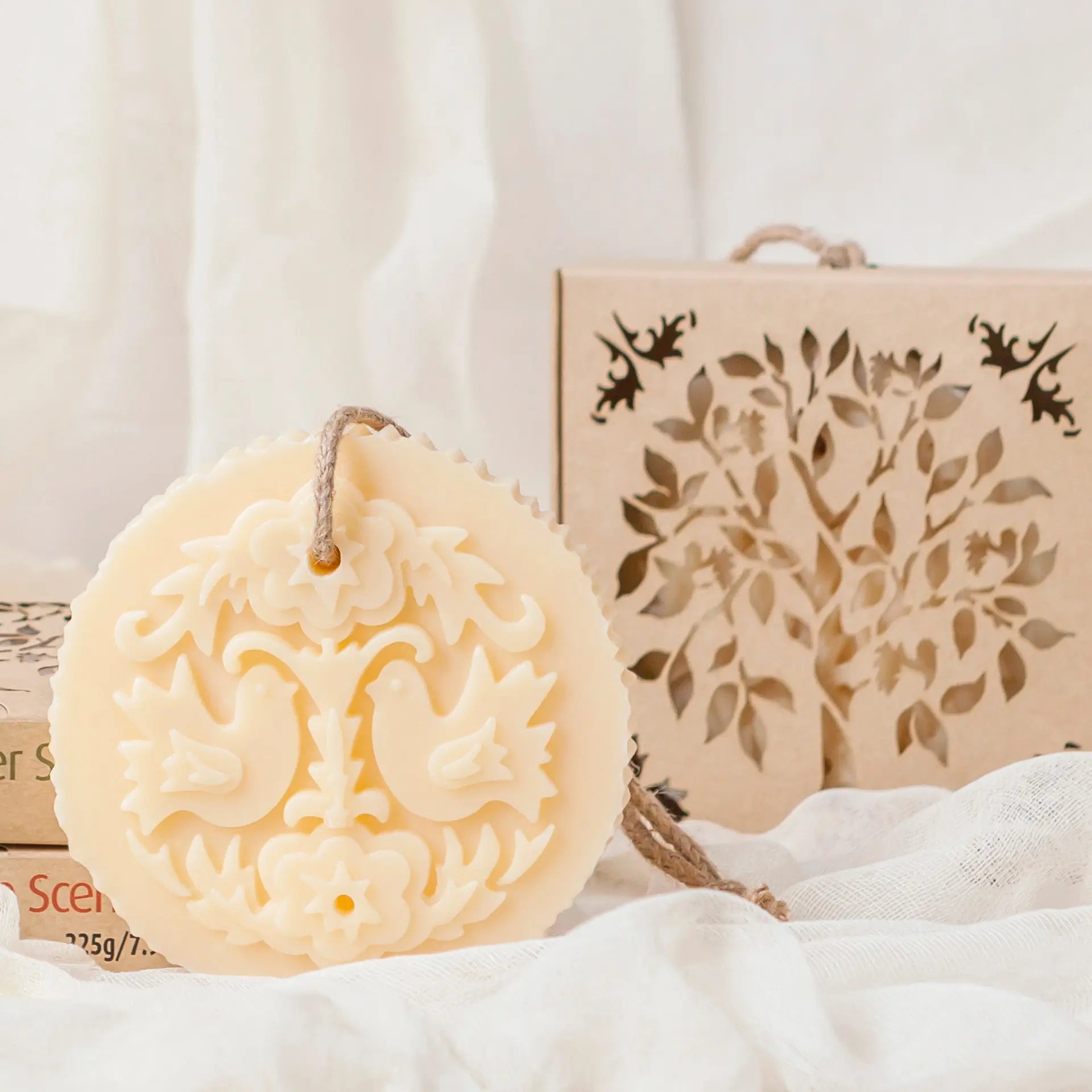 Castelbel Lovebirds Soap on Rope,  round cream-coloured soap bar with an embossed floral lovebird design, hanging from a twine loop, displayed next to a decorative kraft paper gift box with a laser-cut tree design.