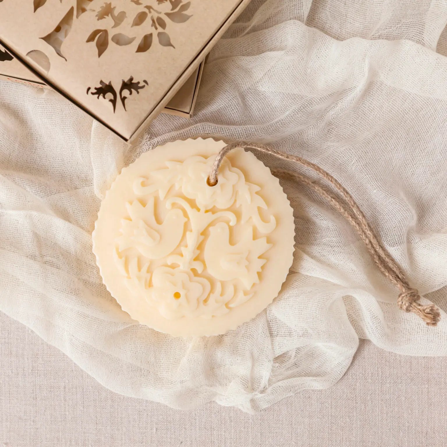 Castelbel Lovebirds Cedar & Rose Soap on Rope, round cream-coloured milled soap bar on draped white fabric next to a wooden box with laser-cut leaf details