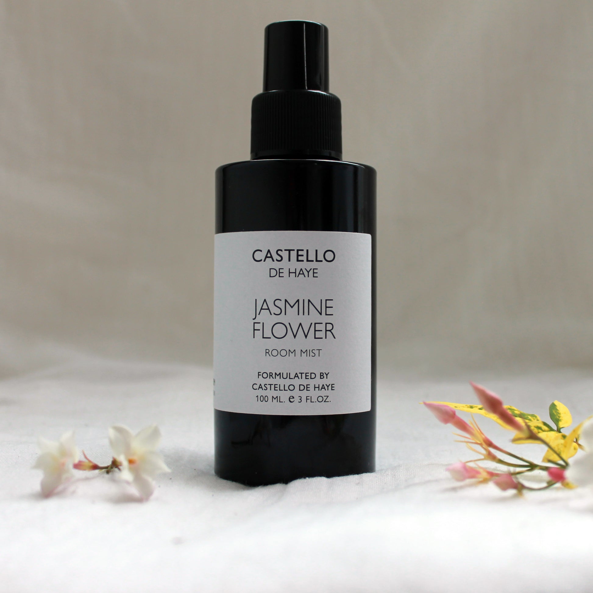 A black spray bottle labeled 'Castello de Haye Jasmine Flower Room Mist' with violet glass and flowers around it.