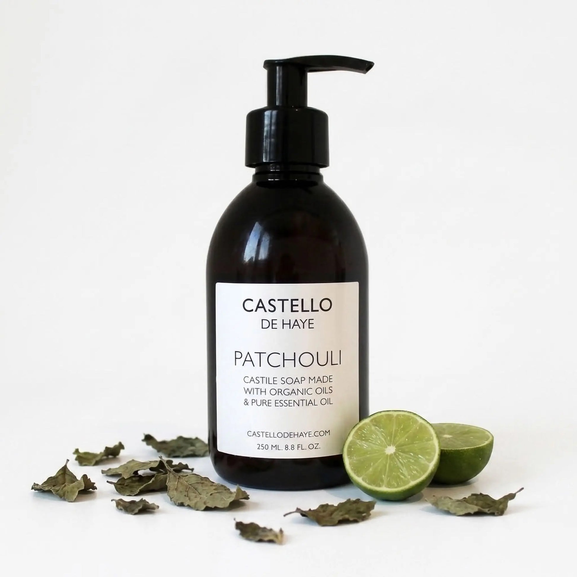 Castello de Haye Patchouli and lime hand and body Castile soap in a dark amber bottle with a black pump, displayed on a white background with dried patchouli leaves and sliced lime