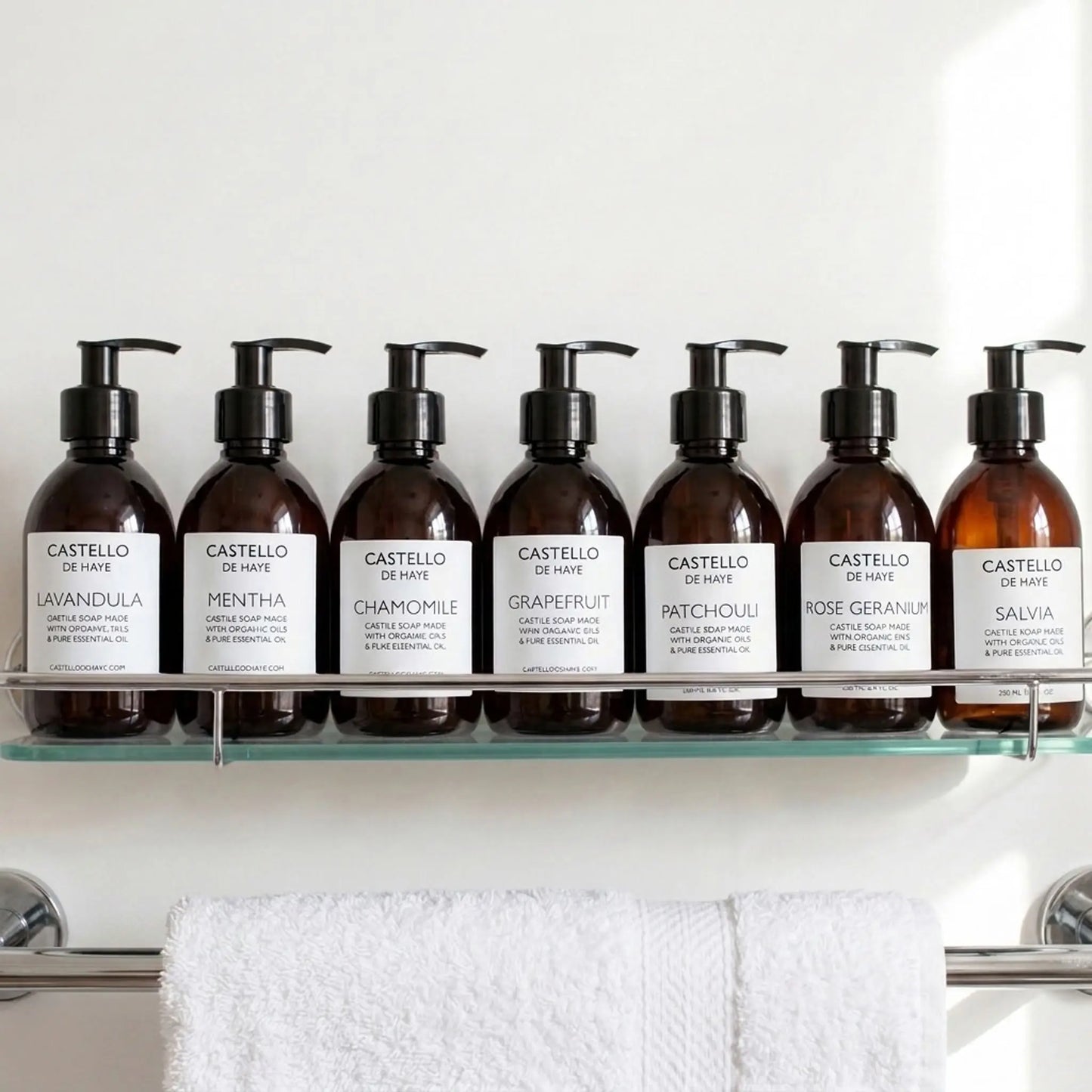 Six amber pump bottles of Castello de Haye Castile Soap arranged on a clear glass shelf above a white towel. The bottles have black pumps and white labels showing scents: Lavandula, Mentha, Chamomile, Grapefruit, Patchouli, Rose Geranium, and Salvia.