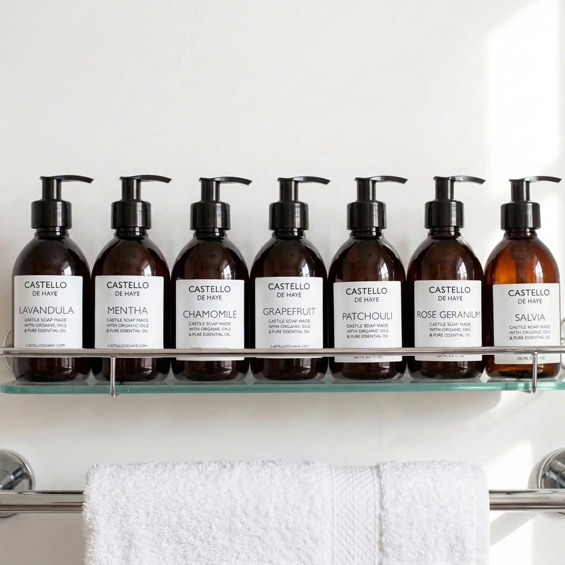 Six amber pump bottles of Castello de Haye Castile Soap arranged on a clear glass shelf above a white towel. The bottles have black pumps and white labels showing scents: Lavandula, Mentha, Chamomile, Grapefruit, Patchouli, Rose Geranium, and Salvia.