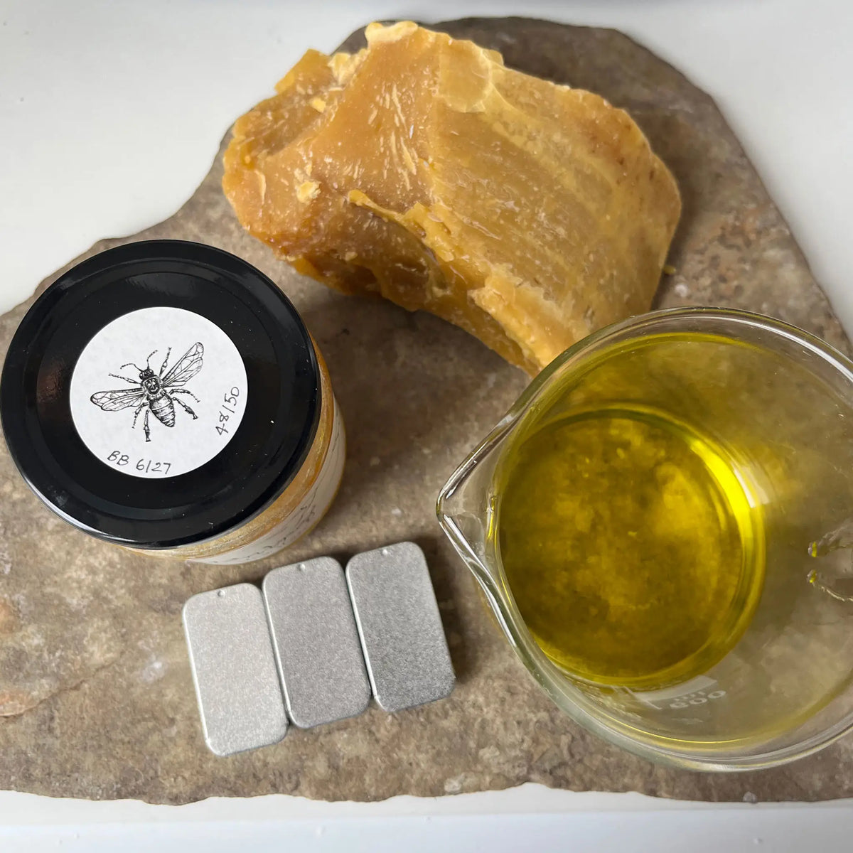 Ingredients for Castello de Haye Lip Balm, Local Beeswax, jar of local honey, and a measuring cup of oils on a stone surface.