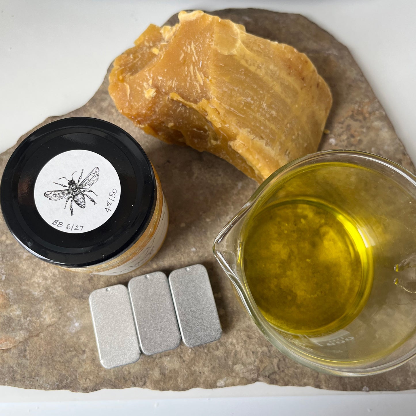 Beeswax, a container with a label, and a measuring cup of liquid on a stone surface.
