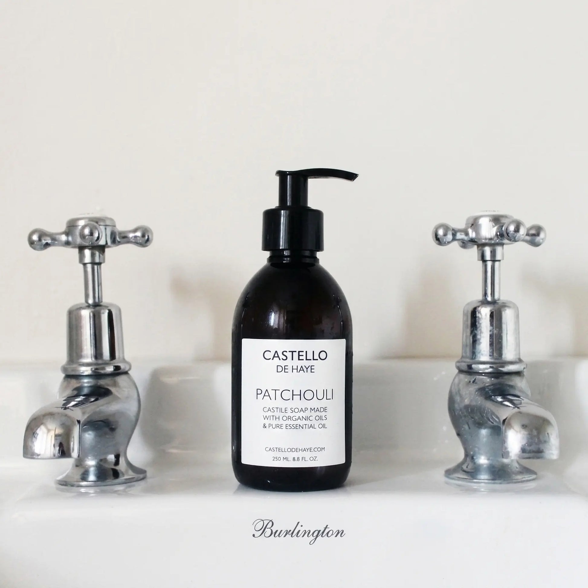 Castello de Haye Patchouli and lime hand and body Castile soap in a dark amber bottle with a black pump, displayed on a Burlington sink ledge between two silver cross-handle taps.