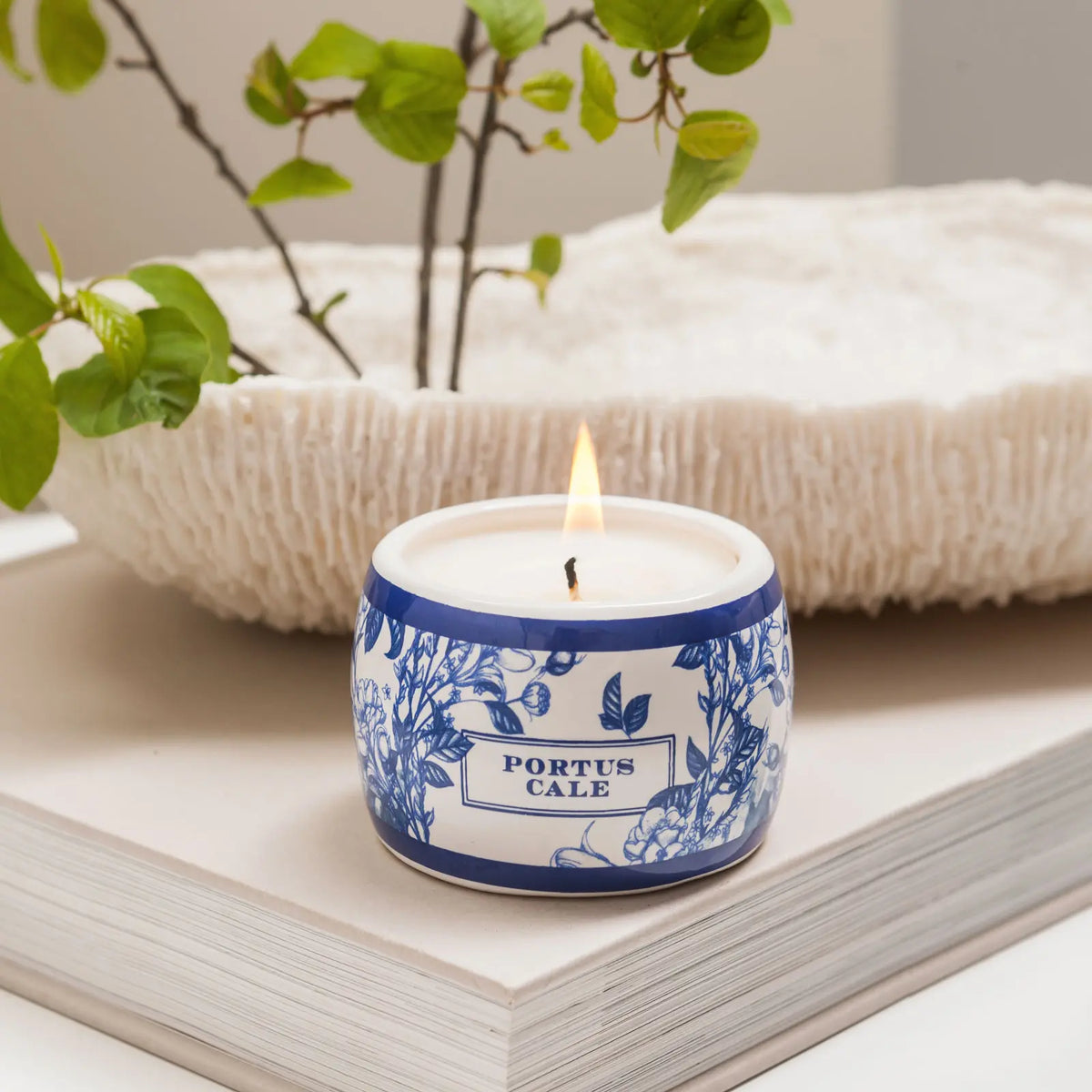 Lit Portus Cale Gold & Blue candle in a patterned ceramic holder, styled on an open book