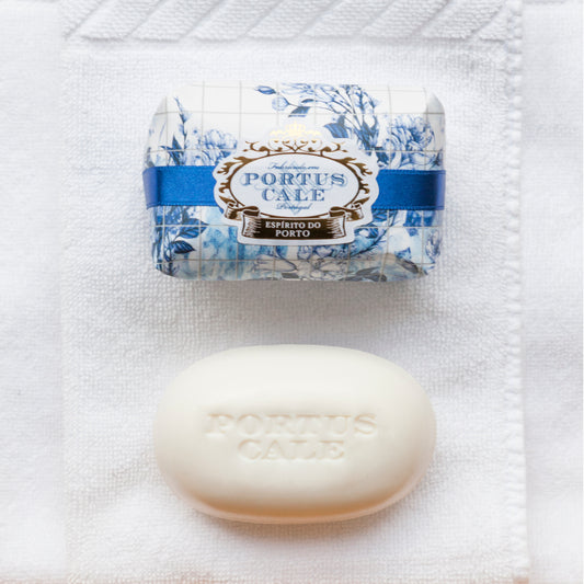 A bar of Portus Cale Gold & Blue soap with a floral design on the wrapping, alongside a plain white oval-shaped stamped soap.