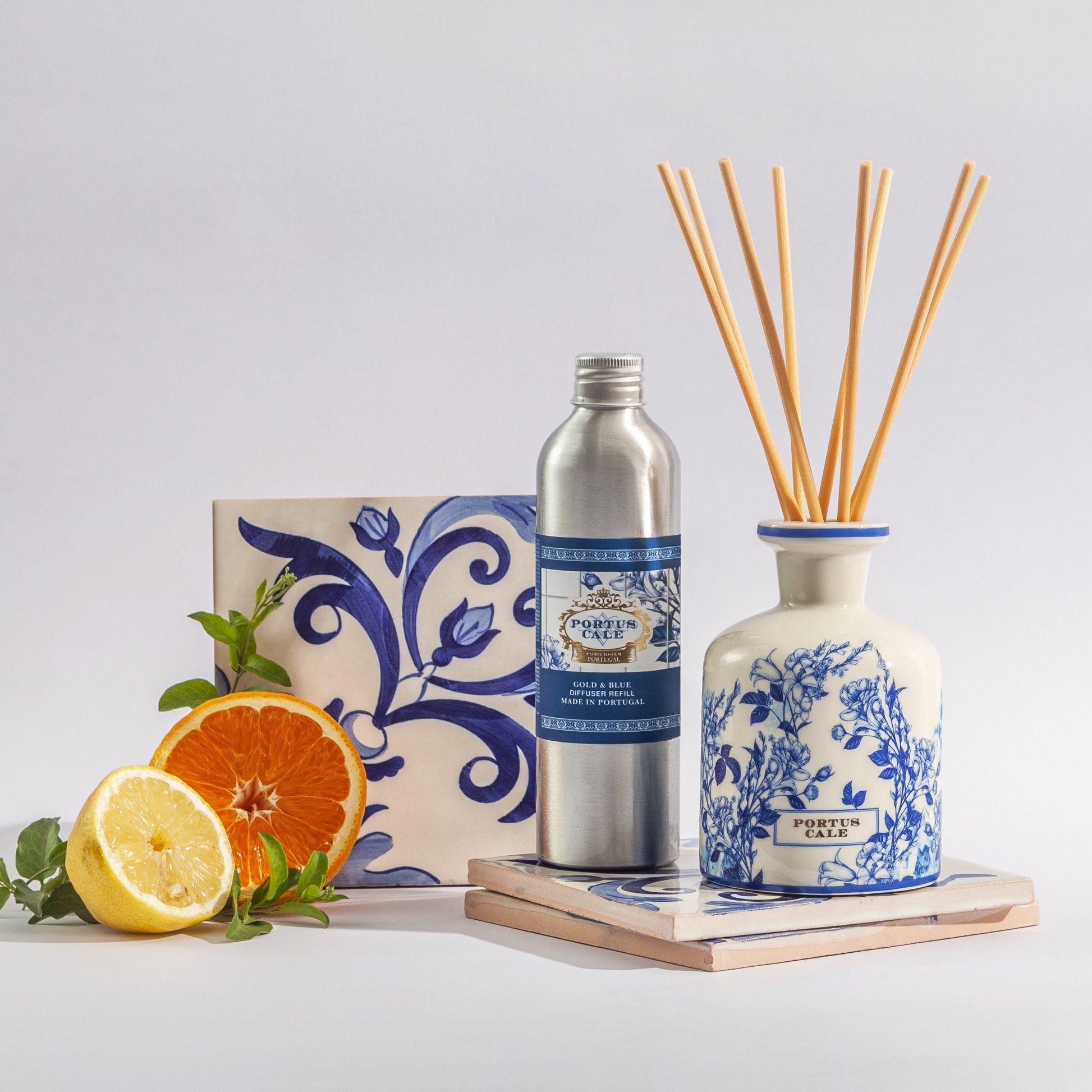 Portus Cale Gold and blue diffuser in a ceramic vase sitting on a traditional portuguese tile, with a bottle of refill next to it and some oranges and lemons cut in half by the side