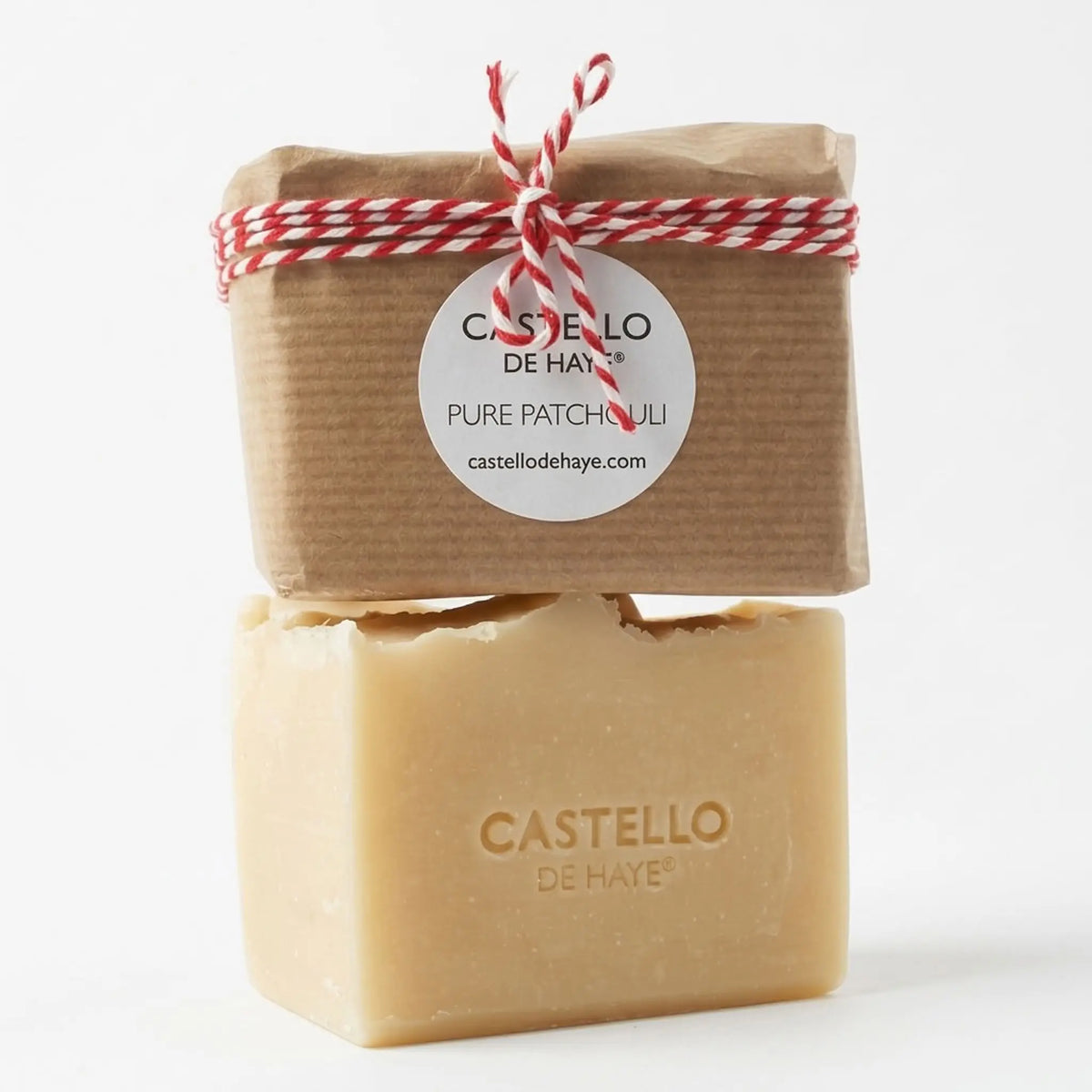 Two stacked bars of Castello de Haye Pure Patchouli soap on a white background, with the top bar wrapped in brown kraft paper and tied with a red and white striped string, and the bottom bar showing the natural beige soap color and embossed brand name.