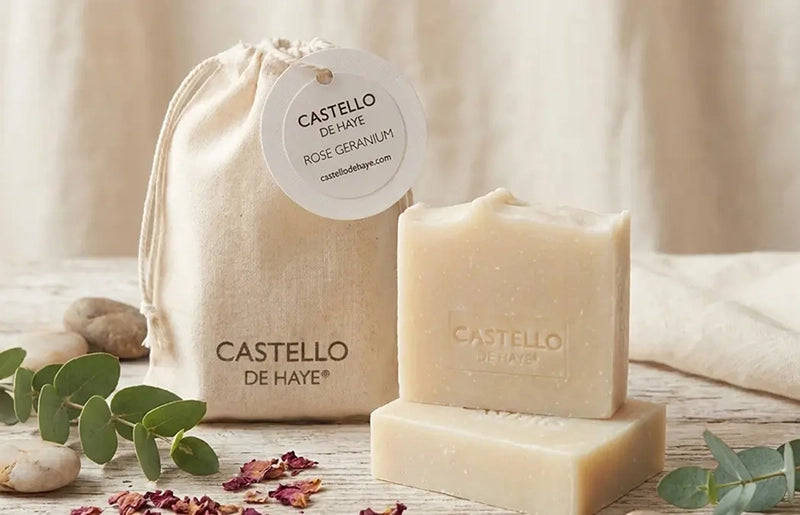 Two bars of Castello de Haye soap with a muslin bag and rose petal and eucalyptus leaves on a neutral background