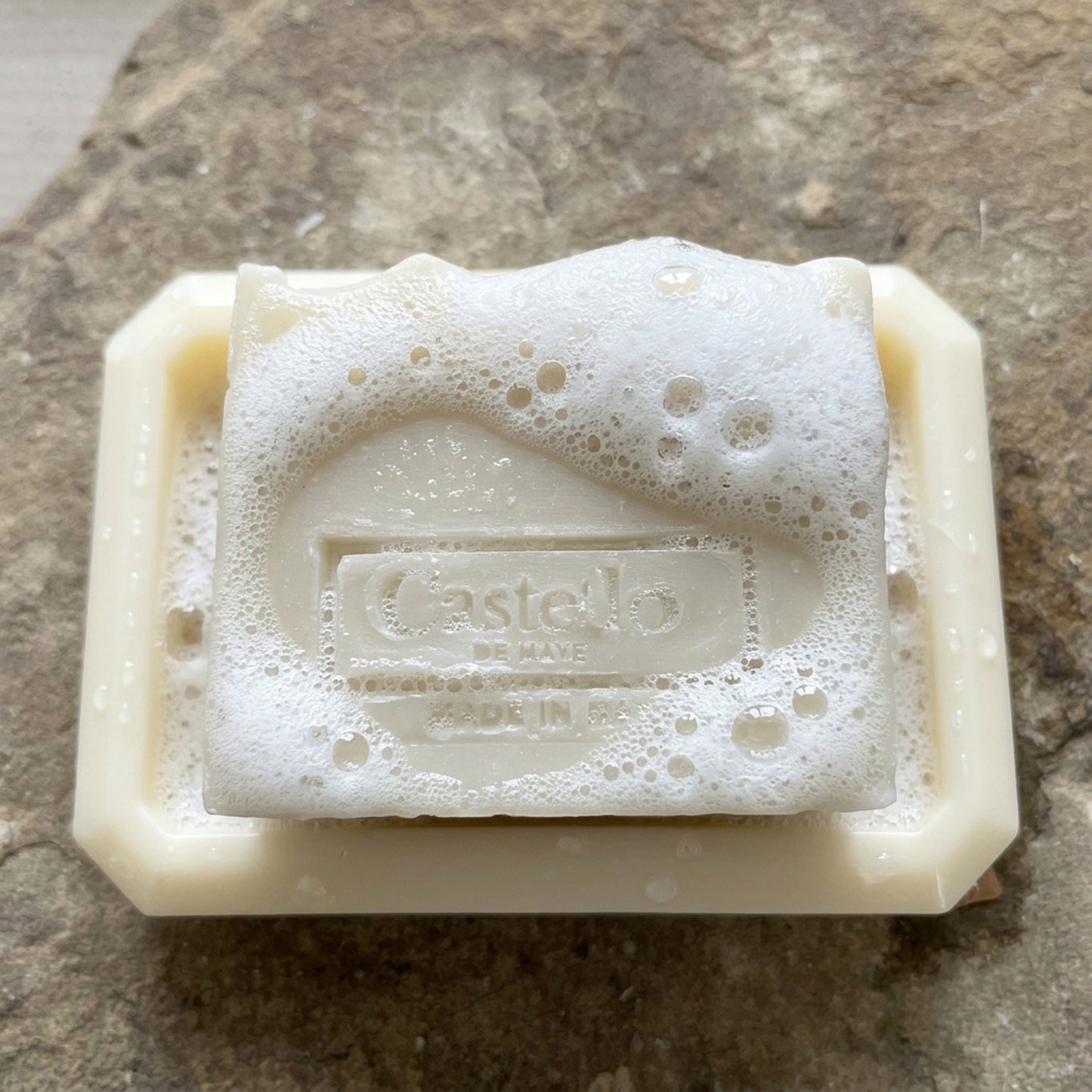 Castello de Haye handmade soap lathering on a white soy wax oblong soap dish on a stone surface.