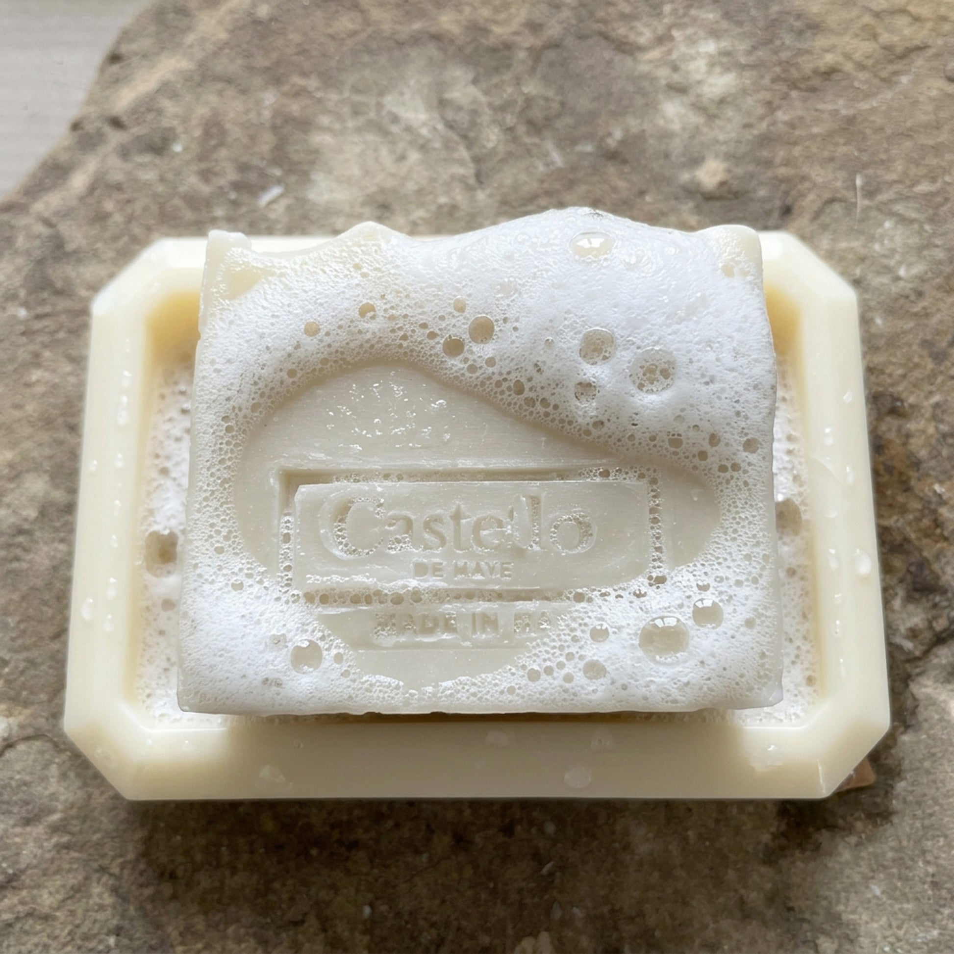 Castello de Haye handmade soap lathering on a white soy wax oblong soap dish on a stone surface.