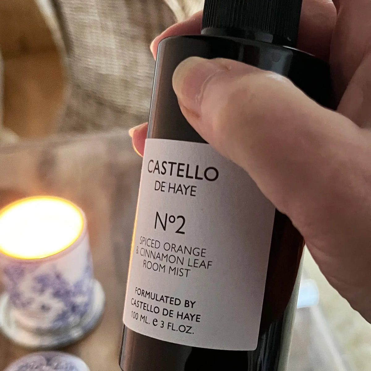 Hand holding Castello de Haye No.2 Spiced Orange & Cinnamon Leaf room mist with a lit candle in the background.