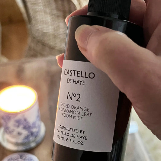 Hand holding Castello de Haye No.2 Spiced Orange & Cinnamon Leaf room mist with a lit candle in the background.