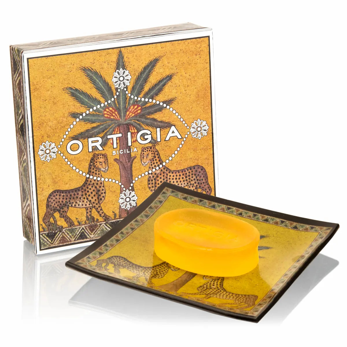 A bar of yellow soap resting on a decorative square dish with a matching branded box behind it. The packaging and dish feature an illustrated design with cheetahs, a palm tree, and the text 'ORTIGIA SICILIA'.