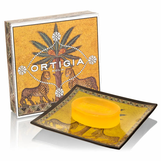 A bar of yellow soap resting on a decorative square dish with a matching branded box behind it. The packaging and dish feature an illustrated design with cheetahs, a palm tree, and the text 'ORTIGIA SICILIA'.